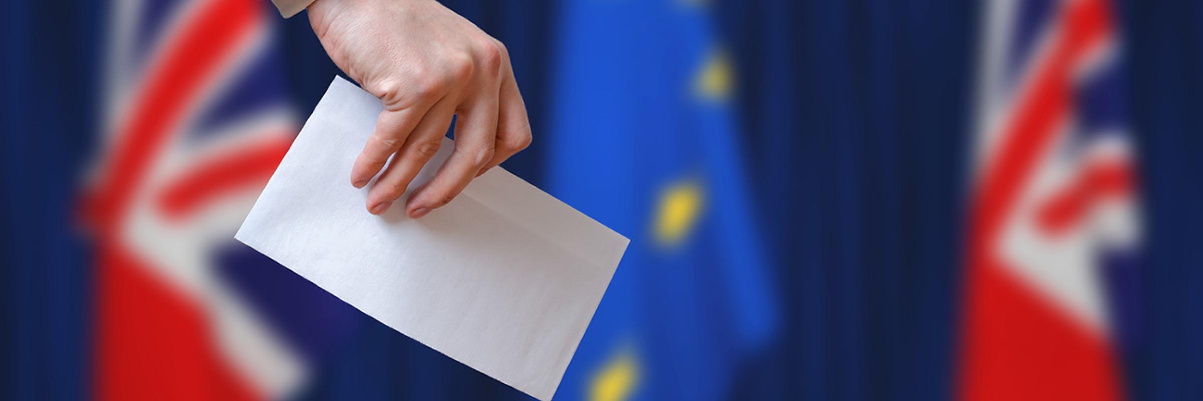 Someone posting a ballot paper in a ballot box, with the Union Jack and E.U flags in the background