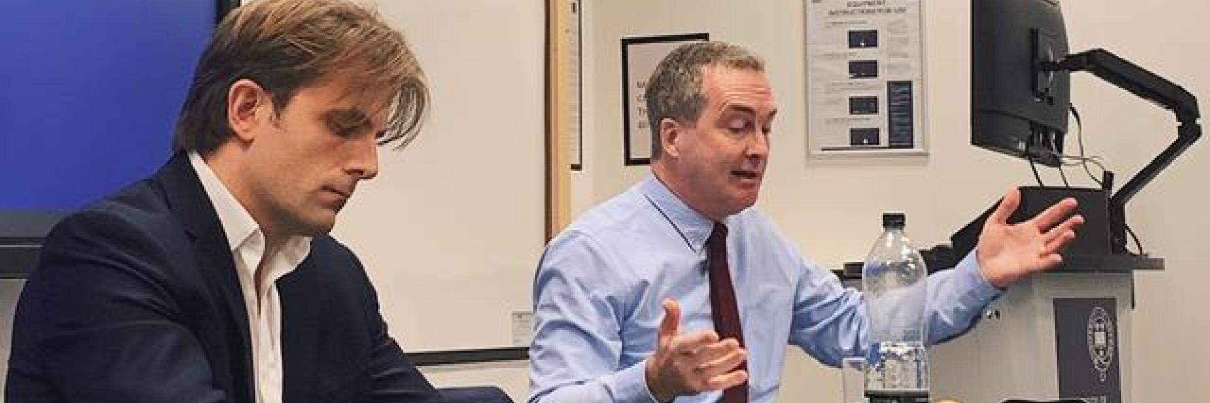 Two men sat at a table presenting in front of an audience