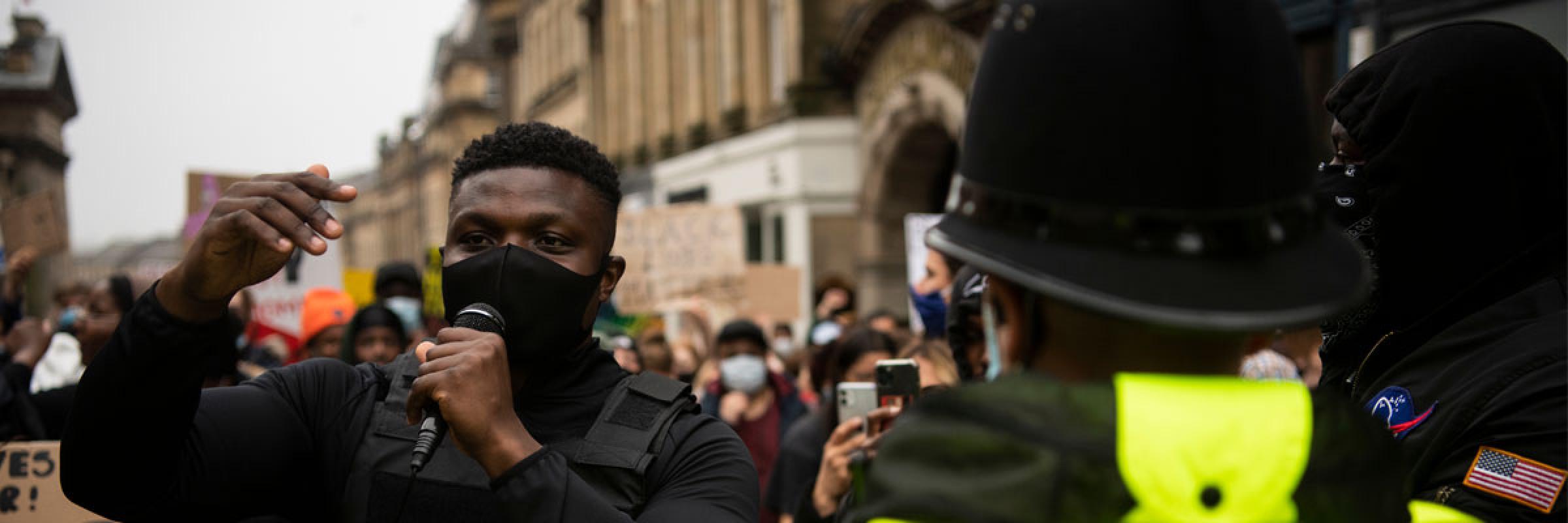 Image of a black man facing camera with a microphone in his hand with a policeman facing him