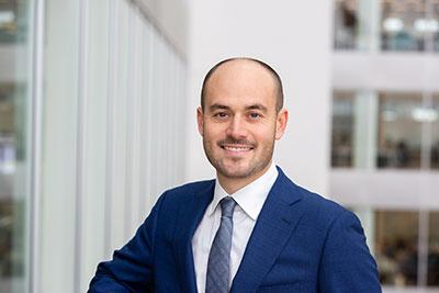 Image of a man smiling at camera wearing a blue suit and tie with a building behind him