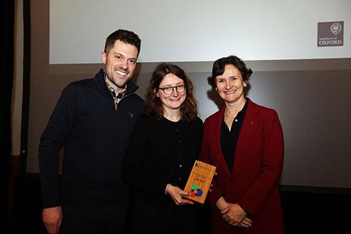 Image of a woman holding a certificate stood in front of a wall with a man on one side of her and a woman on the other side of her, with the University of Oxford logo on the wall