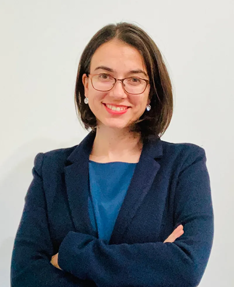 Image of a woman wearing glasses, a dark blue jacket and blue top smiling at camera